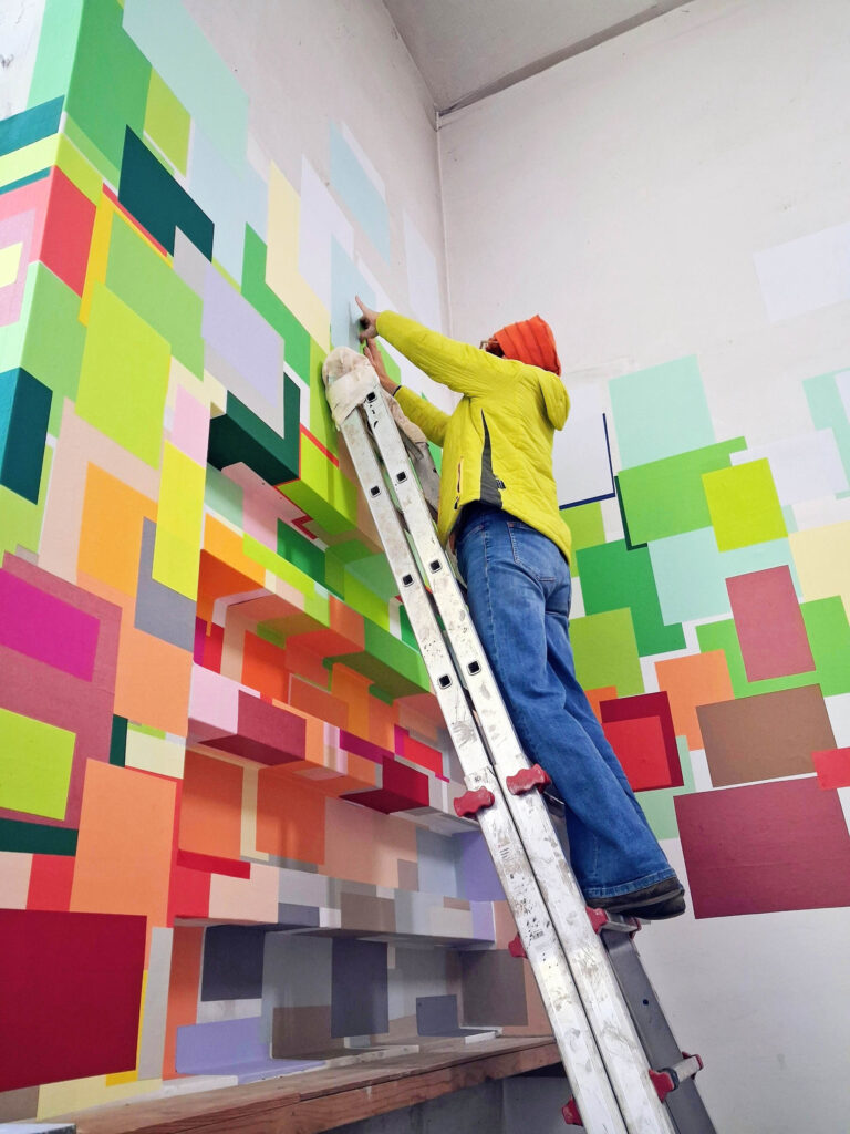 Annegret Hoch auf einer Leiter beim Installieren einer raumgreifenden Arbeit mit bunten Rechtecken an der Wand, in Seitenansicht